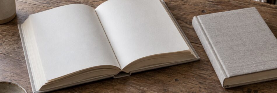 An open blank book on a wooden table beside a pencil